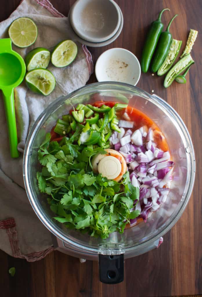 Bowl of food processor filled with fire roasted tomatoes, serrano chiles, chopped red onion, fresh cilantro.