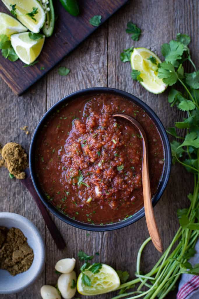 Bowl of canned tomato salsa with a wooden spoon on top. Side of limes, cilantro, and cumin.