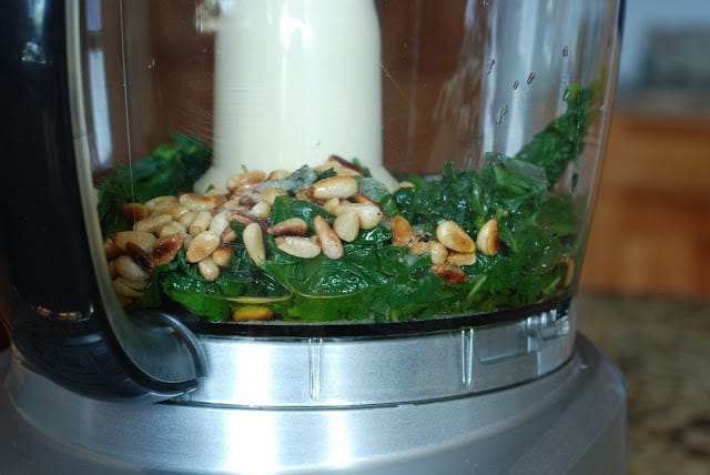 food processor bowl filled with blanched basil, pinenuts, olive oil