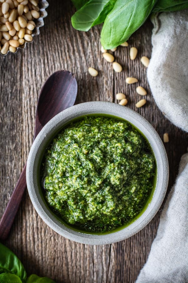 gray bowl filled with perfect basil pesto on a rustic wood board with pine nuts and basil around edges