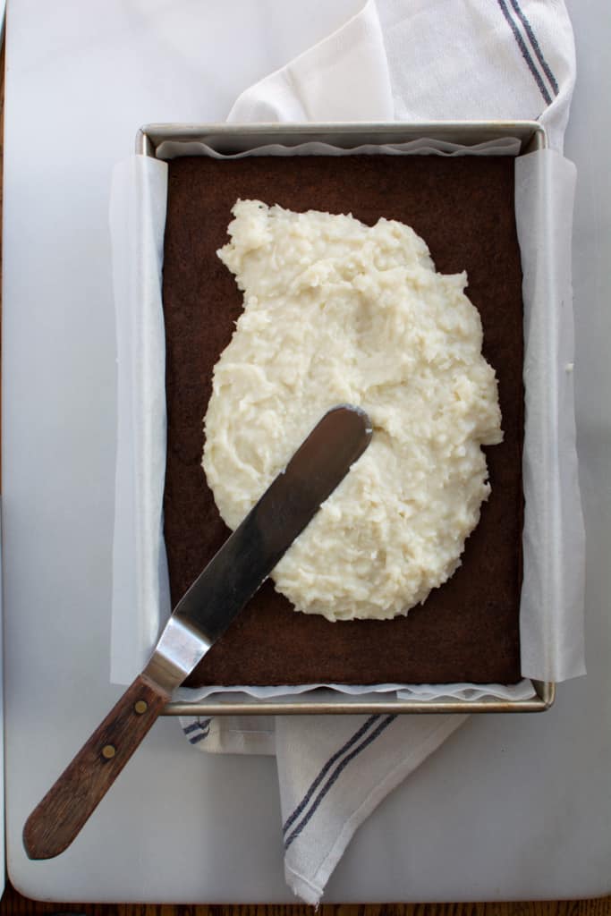Rectangular pan of baked brownies with coconut cream filling spread on top with metal spatula.