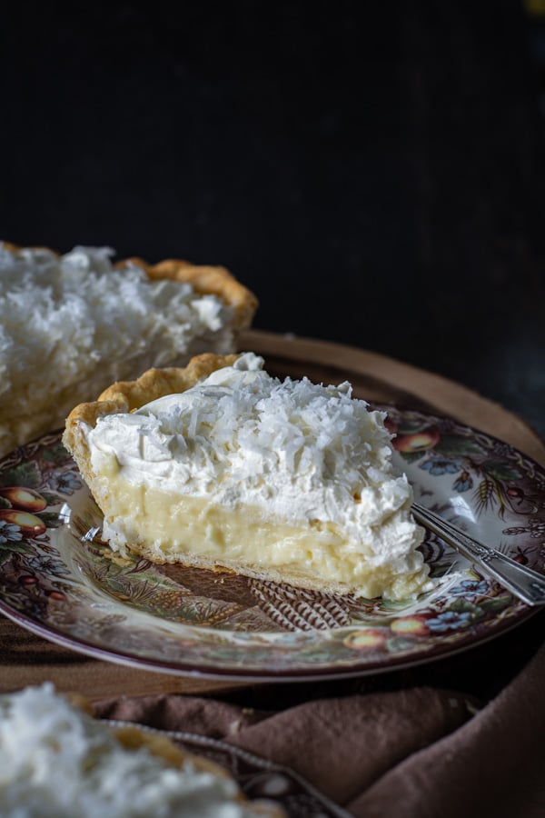 Side view of a slice of coconut cream pie with whole pie in back ground
