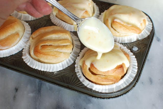 Metal spoon drizzling orange glaze over the baked butterflake rolls.