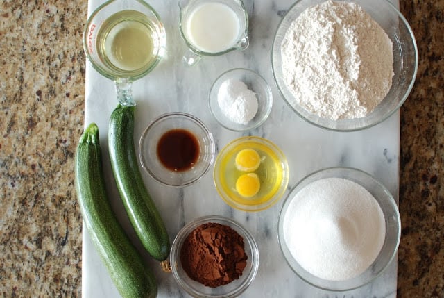 Cake ingredients measured into bowls, 2 zucchini, 2 eggs in bowl
