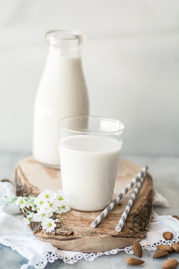 Glass of almond milk with 2 gray striped straws on wood board with container of almond milk in background