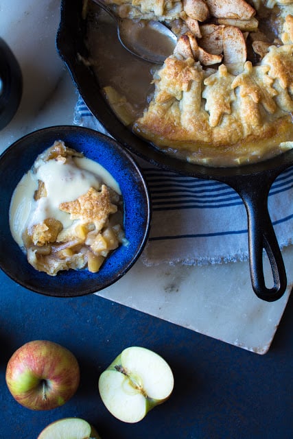 Over head shot apple dumplings in a cast iron pot with a bowl with a drizzle of creme anglaise