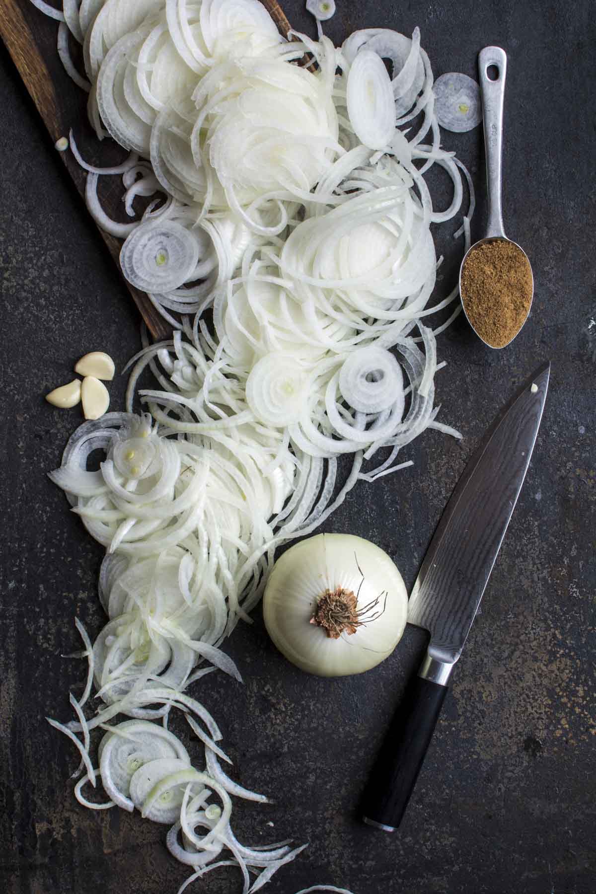 Sliced onions on a board with garlic, knife, and coconut sugar on the side