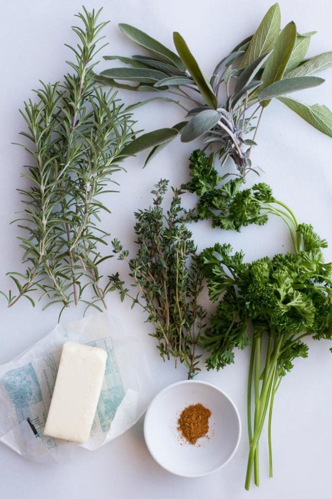 Piles of fresh thyme, sage, parsley, and rosemary. A stick of butter and small white bowl of nutmeg.