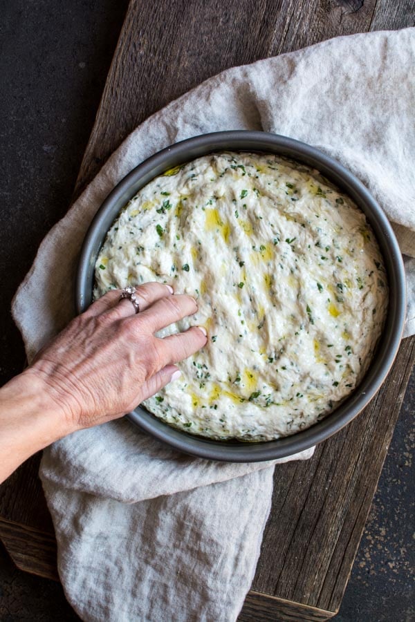 focaccia dough in round baking pan with olive oil on top and fingers pressing into dough