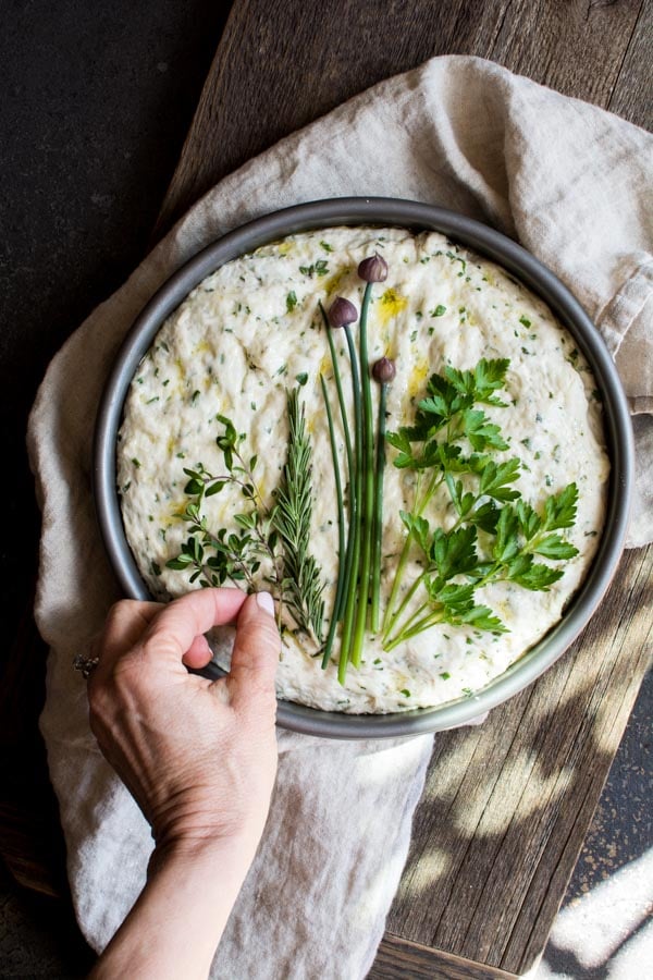 herb focaccia dough in round baking pan topped with arrange fresh herbs