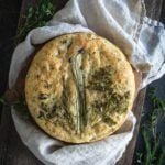 Round focaccia bread topped with an arrangement of fresh herbs on a cloth and bread board