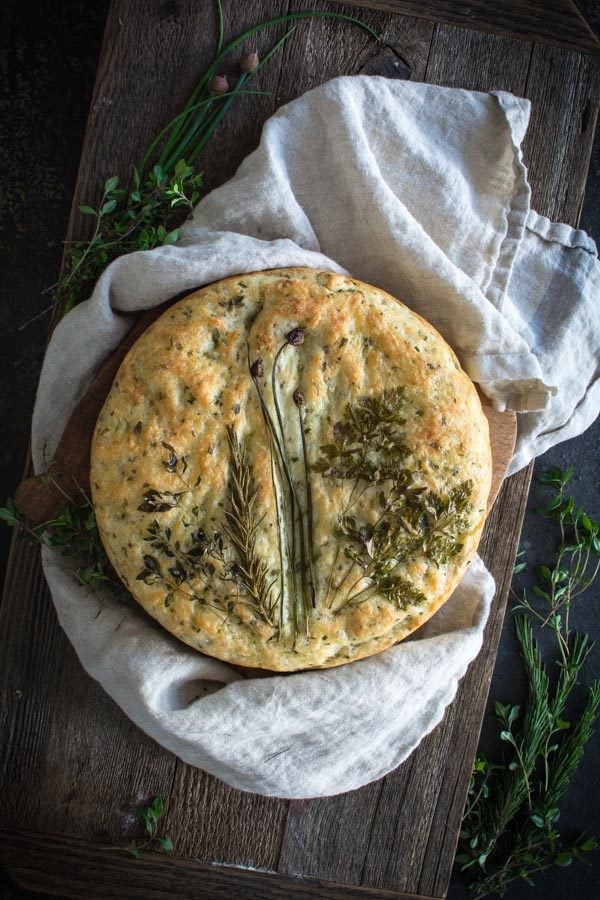 Round focaccia bread topped with an arrangement of fresh herbs on a cloth and bread board