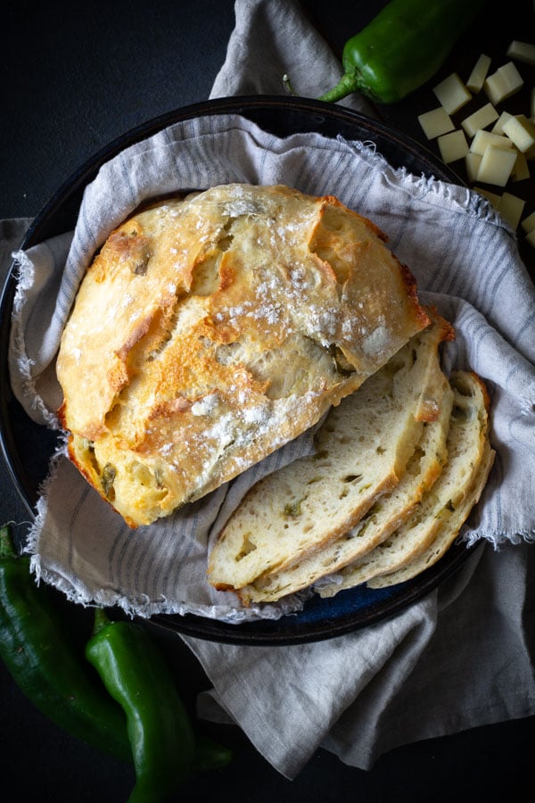 No-knead Green Chile Cheddar Bread slices on linen cloth with green chilis and cheese cubs