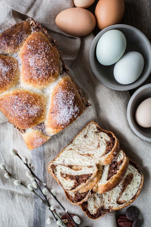 Top down Easter bread with chocolate and pecan swirls. Brioche loaf is cut in half, with one half solid and the other cut into two slices. Gray bowls with pink and blue eggs are to the side for decoration.