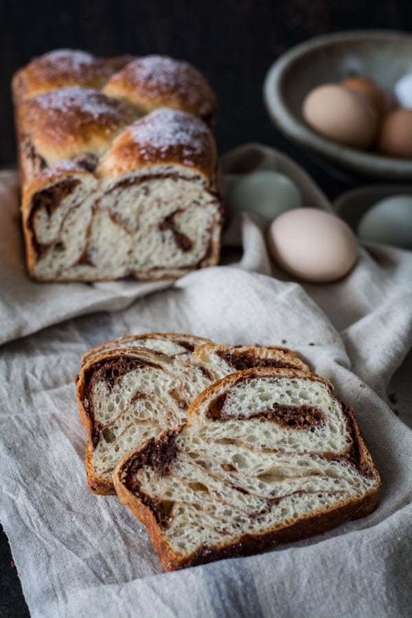 Two slices of no-knead Easter bread with chocolate and pecan swirls, laying together on a white napkin. Remaining half loaf of bread is in the background, with pink and blue eggs for decoration.