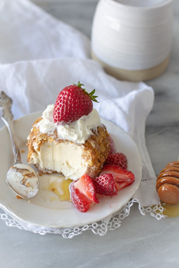 Mexican fried ice cream with strawberries, on a white plate with honey drizzle and honey stick.