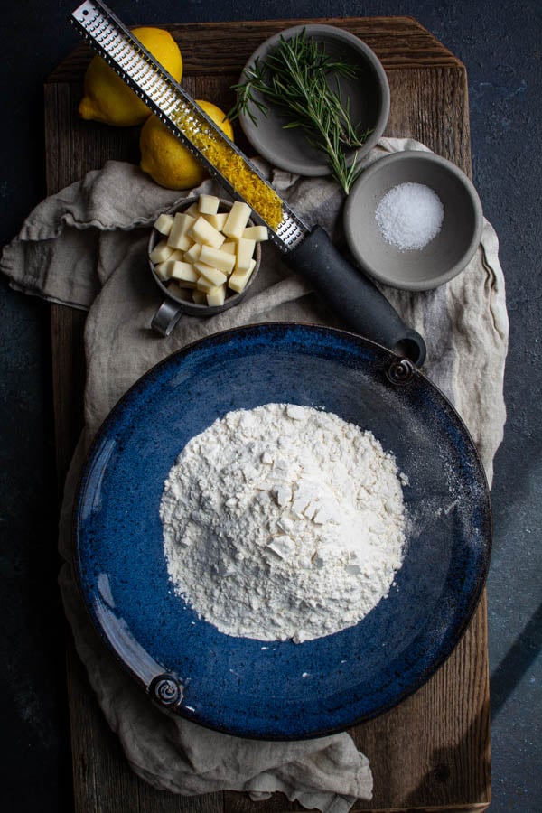 blue bowl with flour surrounded by cubed cheese, lemon zest, rosemary