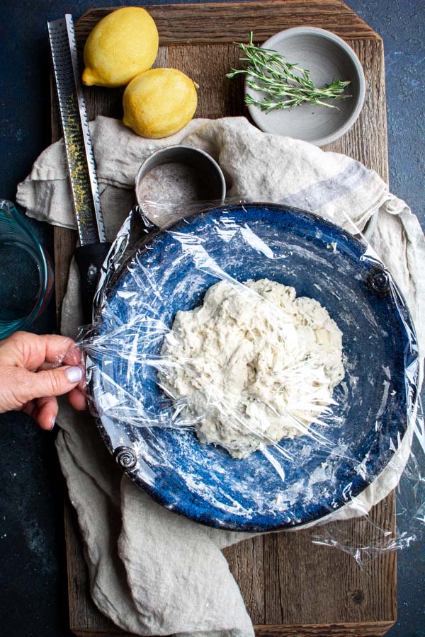 mixed bread dough in blue bowl with plastic wrap on top