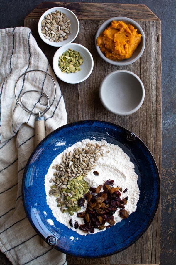 A large blue bowl on a wood board. the bowl is filled with flour topped with dried fruit and seeds.