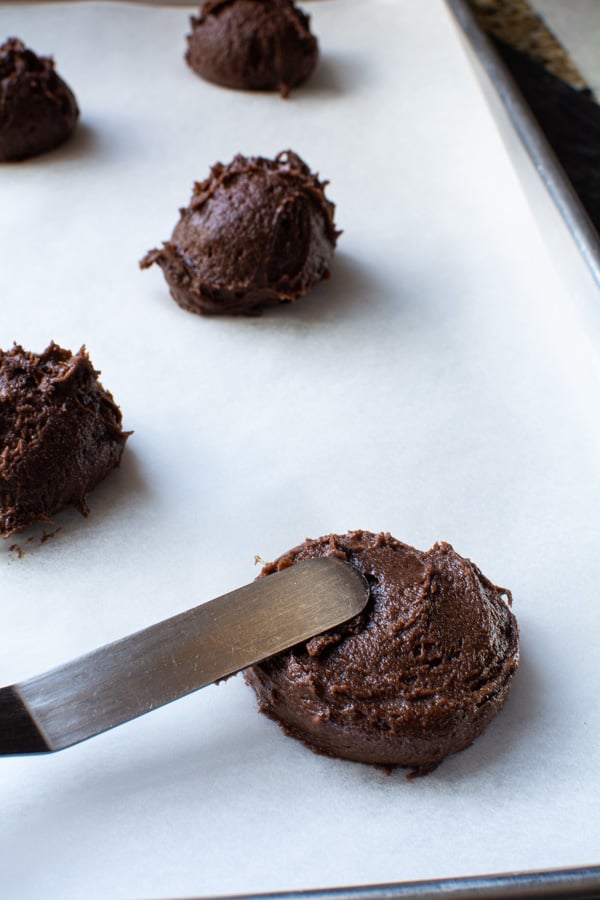 Mounds of chocolate cookie dough on a parchment lined baking sheet spreading with a spatula