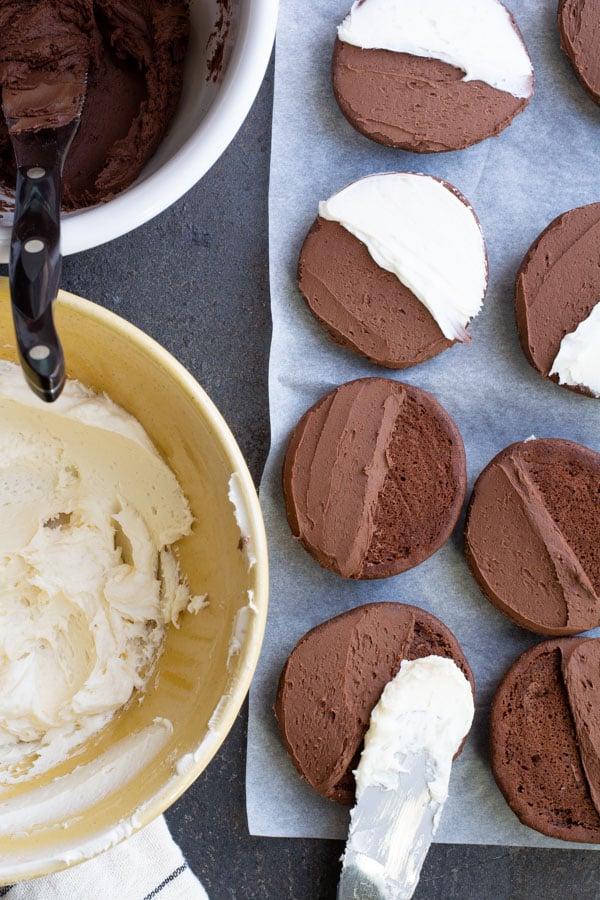 Bowl of vanilla icing and a bowl of chocolate icing along side of cookies partially frosted