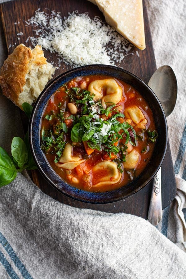 Overhead shot of minestrone soup with cheese tortellini in a bowl with a chunk of bread and fresh grated parmesan cheese.