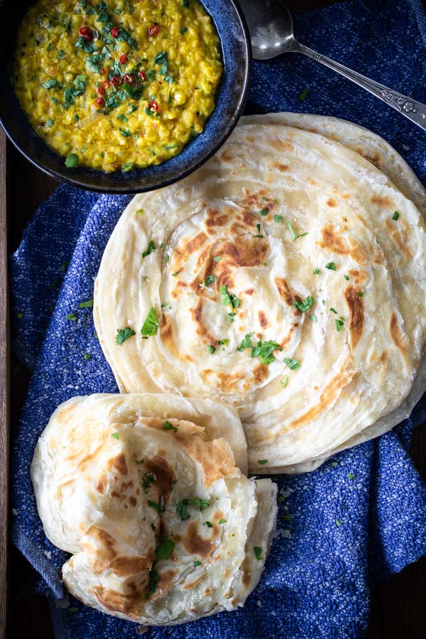 Flakey Paratha flat bread stacked on a cloth with a bowl of lentils