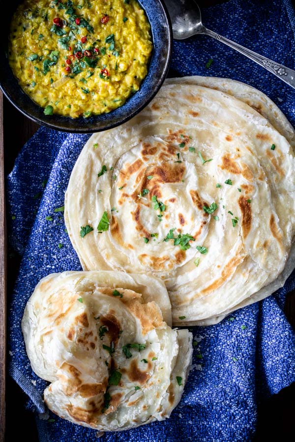 Flakey Paratha flat bread stacked on a cloth with a bowl of lentils