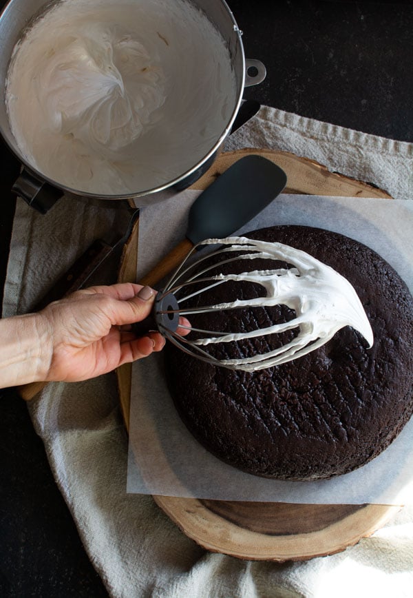 Round chocolate devil's food cake on a wooden board with a whisk of boiled icing dripping off.