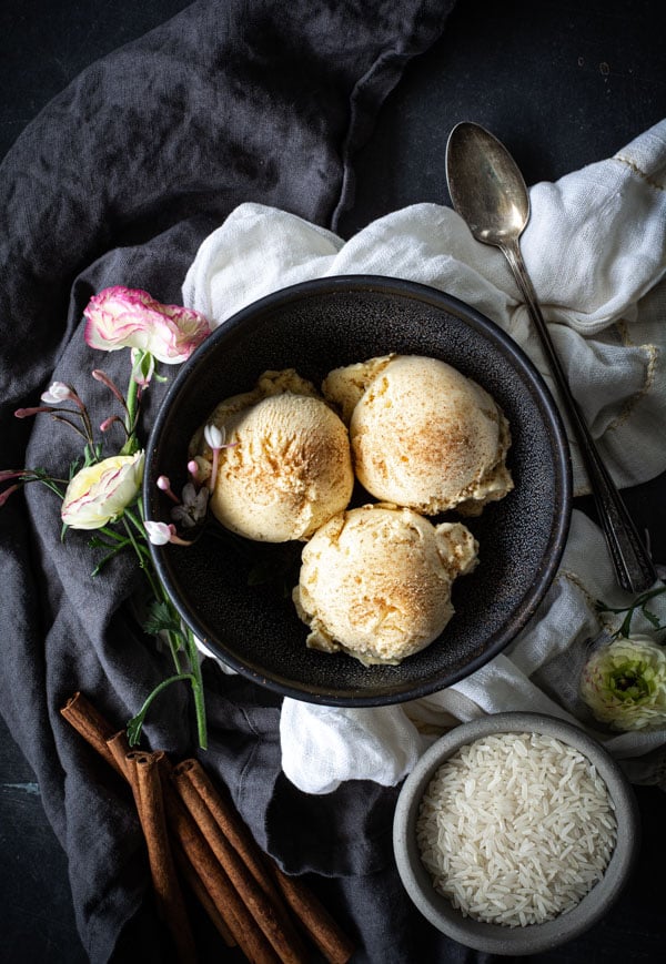 Overhead shot of 3 scoops of Horchata Ice Cream in a bowl, with a bowl of raw rice, cinnamon sticks, and flowers