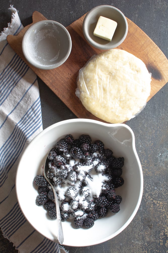 Blackberries with sugar in large bowl with sugar, butter and pie dough on cutting board