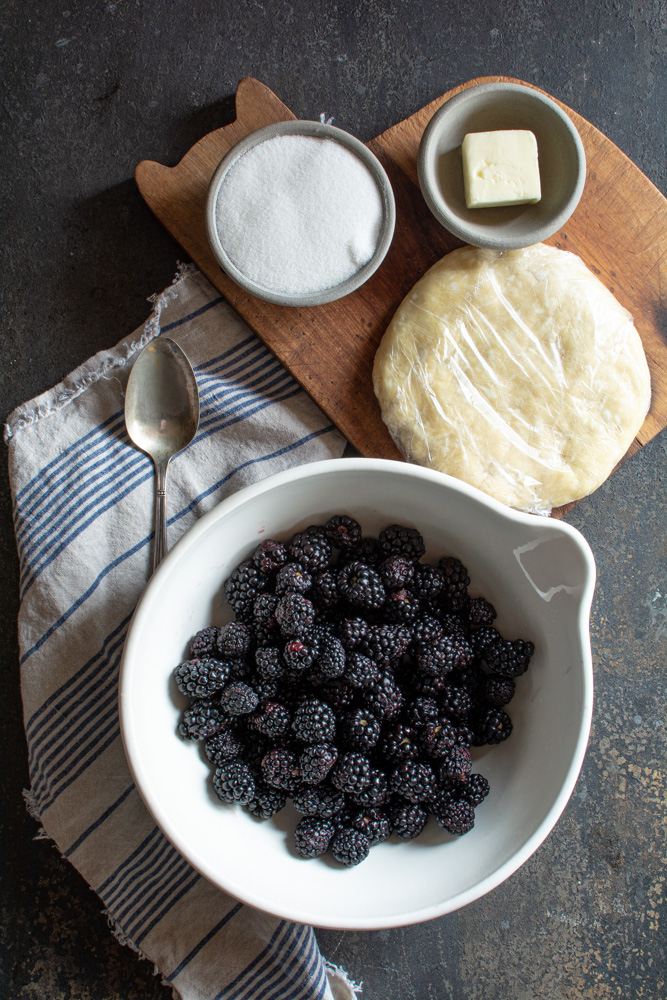 Fresh blackberries in a large bowl with sugar, butter, and pie dough on a board