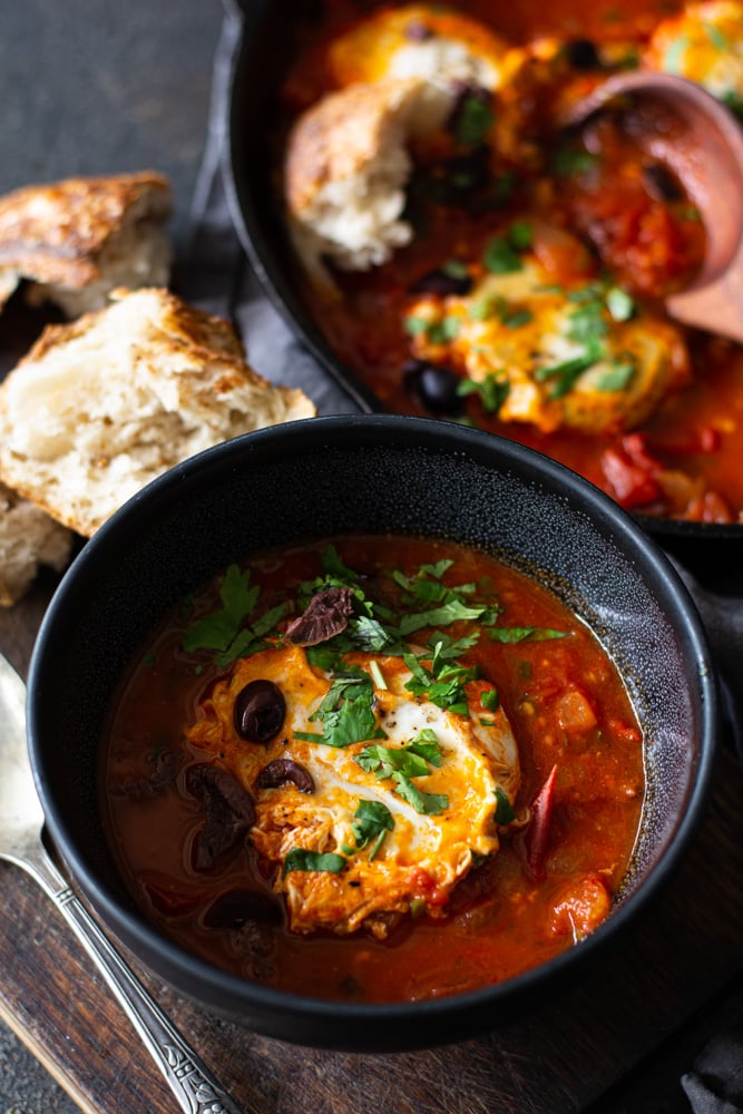 Large bowl full of shakshuka with torn bread and skillet in background