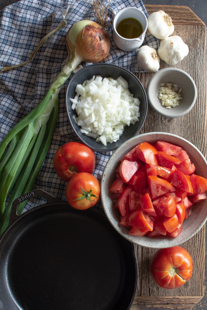 Overhead shot of cast iron skillet, large tomatoe, bowl of chopped tomatoes, onions, garlic