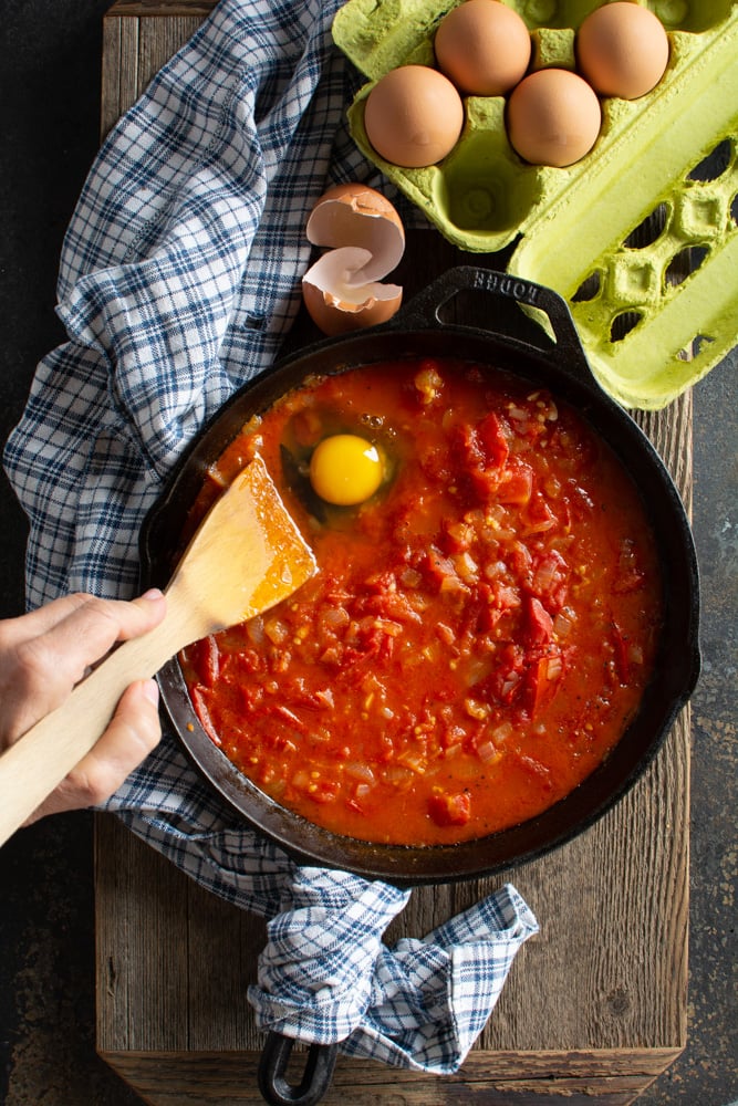 Cooked tomatoes in cast iron skillet with a raw egg dropped on top of sauce.