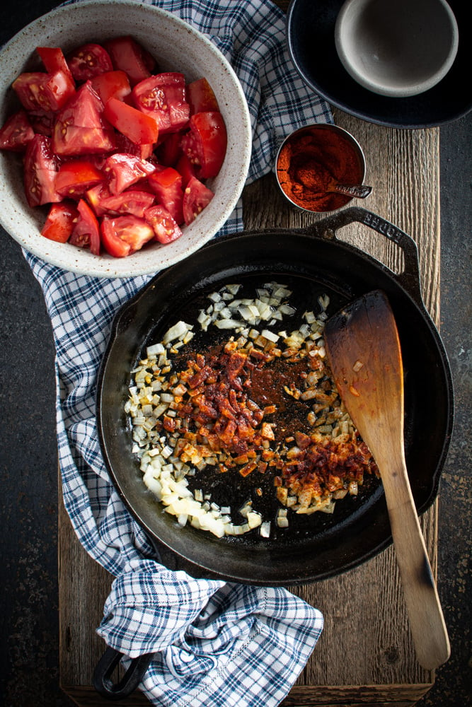 Cast iron skillet with sauteed onions and added smoked paprika. With bowl of chopped tomatoes on the side