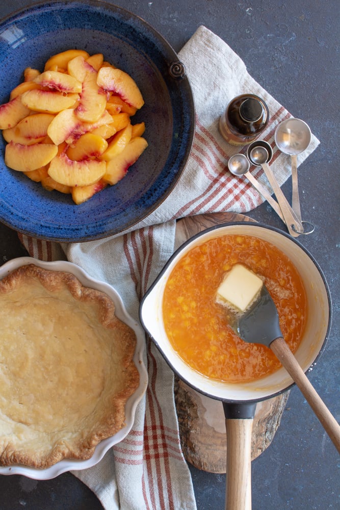 A bowl of fresh sliced peaches, baked pie shell, and a pot with peach glaze with a pat of butter.