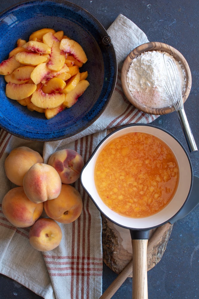 A bowl of sliced peaches, a bowl of sugar and cornstarch mixture, 5 peaches next to a pot of peach glaze.