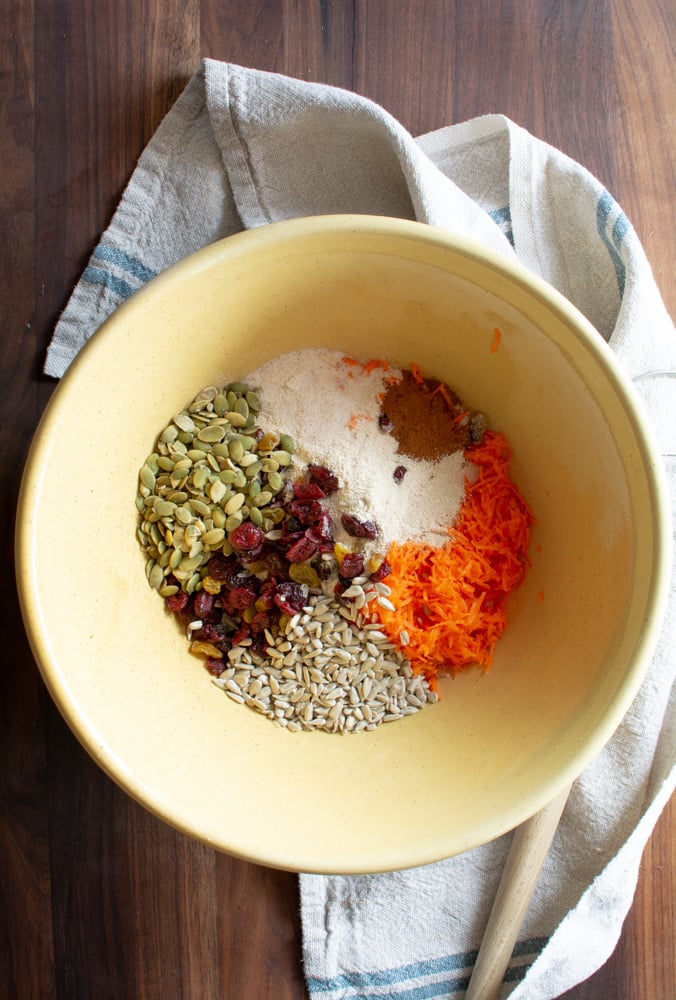 Large yellow bowl filled with seeds, shredded carrots, cinnamon, flour, and dried fruit.