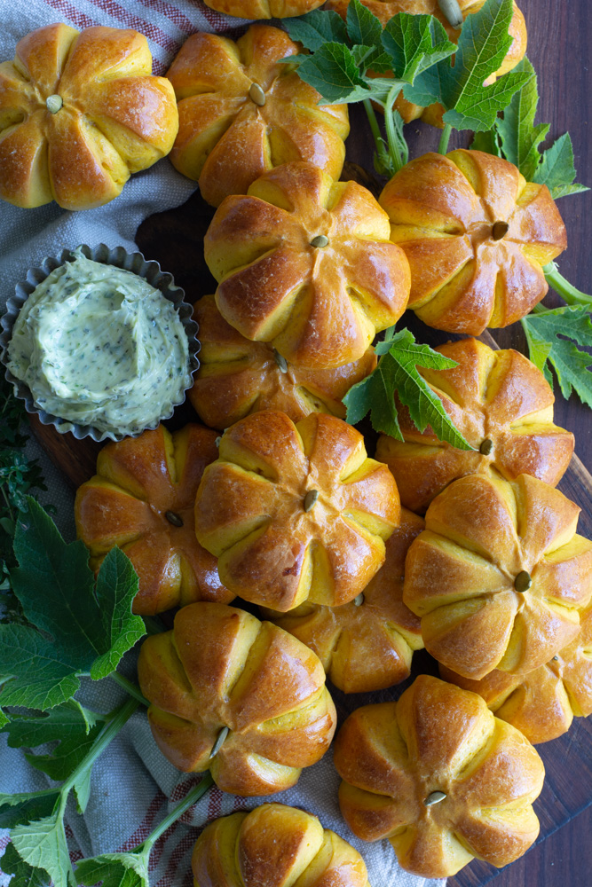 A dozen small pumpkin shaped buns on a platter, surrounded by fresh green leaves and served with a small metal ramekin of homemade herb butter.