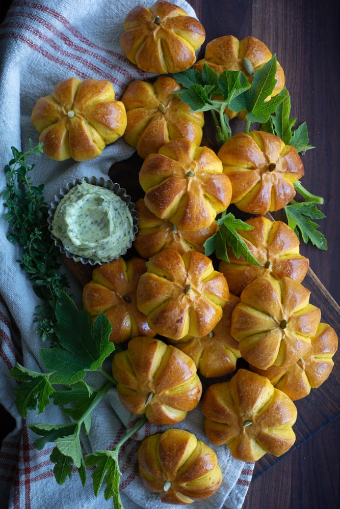 A dozen small pumpkin shaped buns on a platter, surrounded by fresh green leaves and served with a small metal ramekin of homemade herb butter.