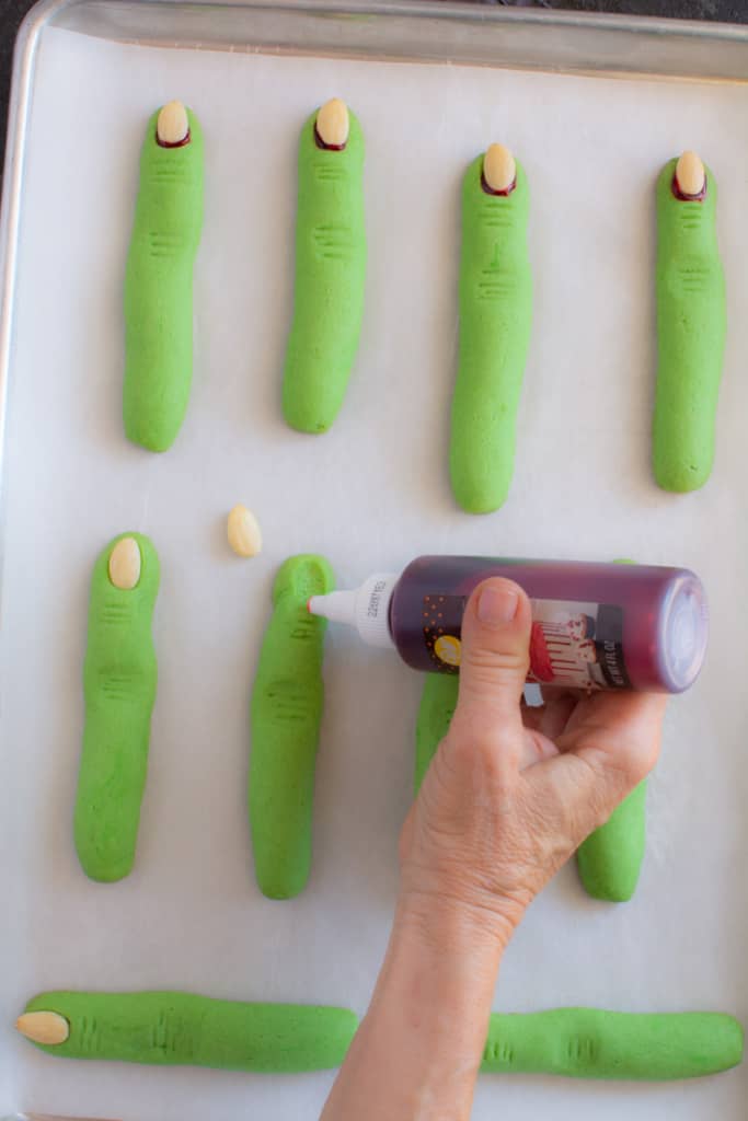 Hand with a squeeze bottle of fake blood, adding to the tips of the green finger-shaped cookies.