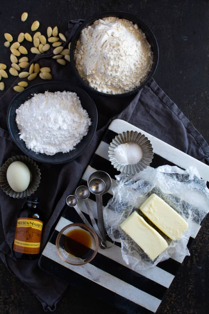 Recipe ingredients top down view: Individual containers of sugar, flour, vanilla, spices, butter, salt, and an egg, Plus silver measuring spoons included.