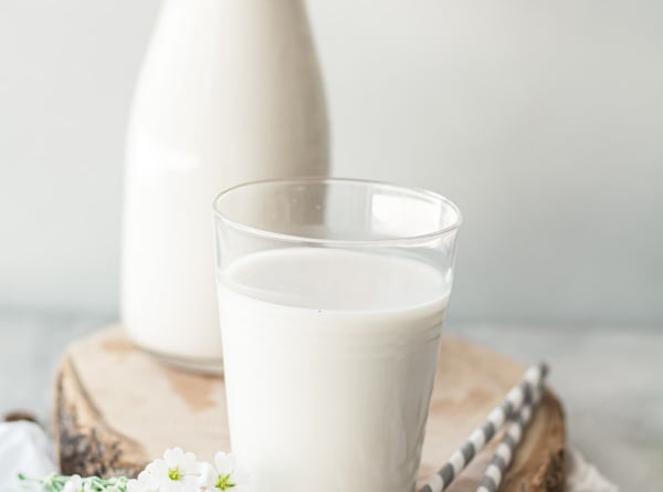 Glass of almond milk with 2 gray striped straws on wood board with container of almond milk in background