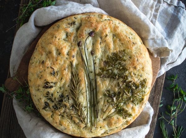Round focaccia bread topped with an arrangement of fresh herbs on a cloth and bread board