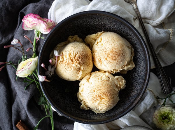 Overhead shot of 3 scoops of Horchata Ice Cream in a bowl, with a bowl of raw rice, cinnamon sticks, and flowers